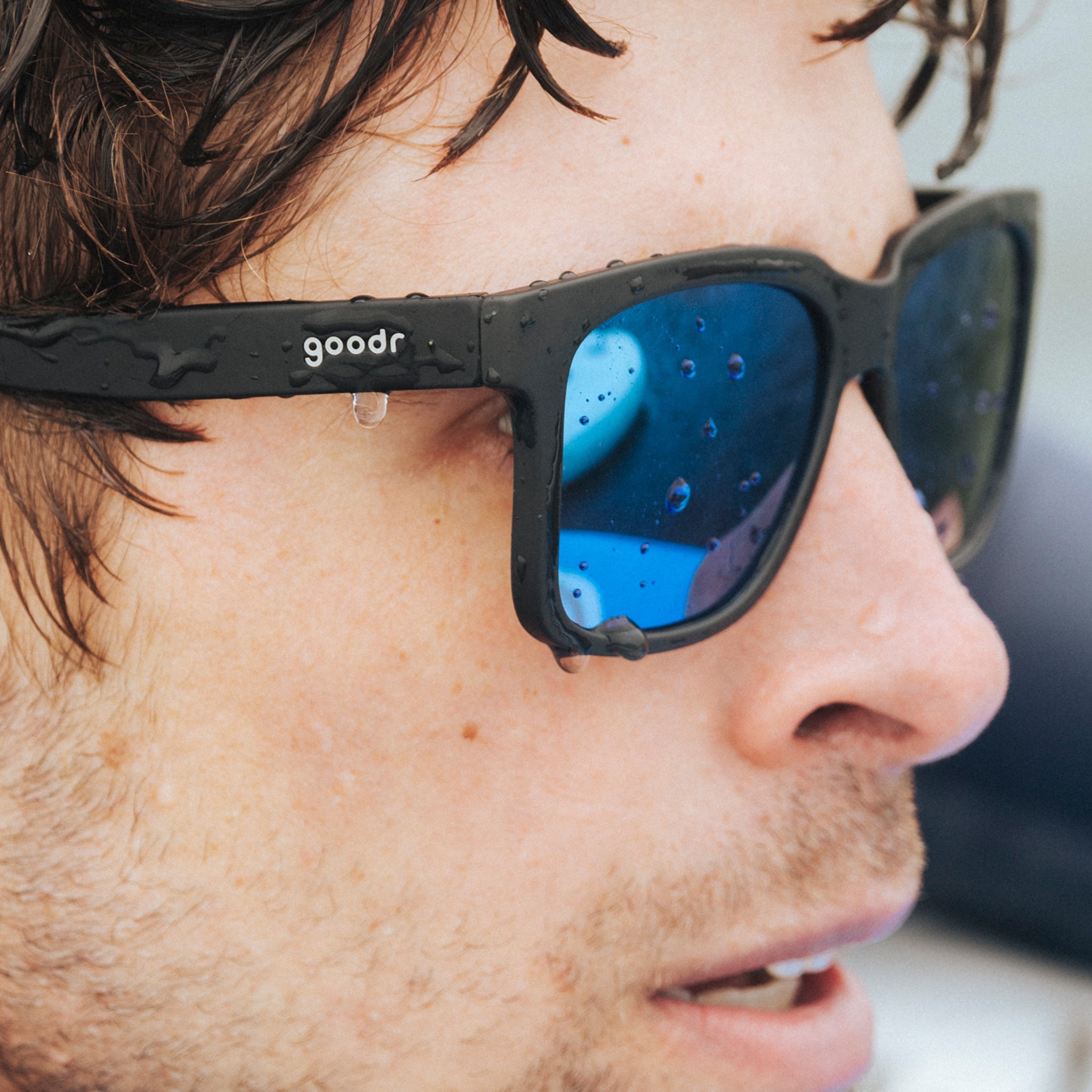 Side view of a young man wearing black sunglasses with blue lenses designed to float in water. Drops roll off the lens.
