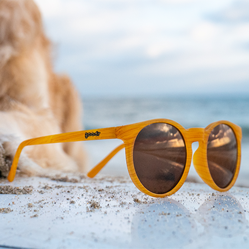 Three-quarter angle view of round brown sunglasses with a wood grain pattern and brown round lenses sunglasses on a beach.