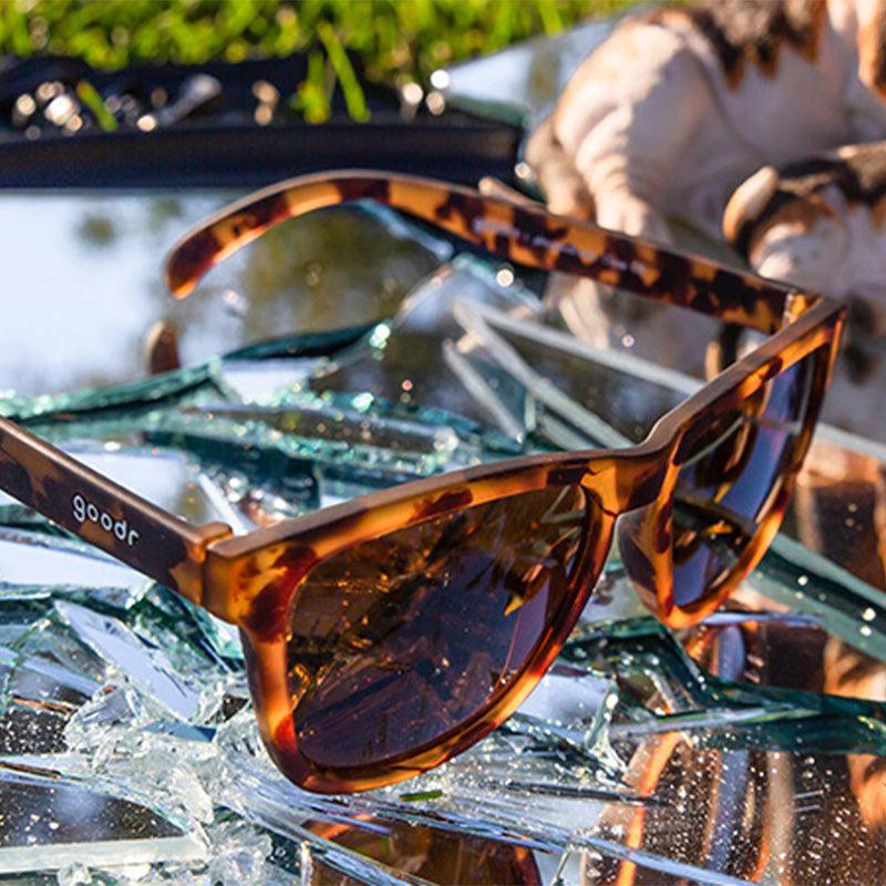 Three-quarter angle view of brown tortoiseshell sunglasses with brown non-reflective lenses sitting atop a broken mirror.