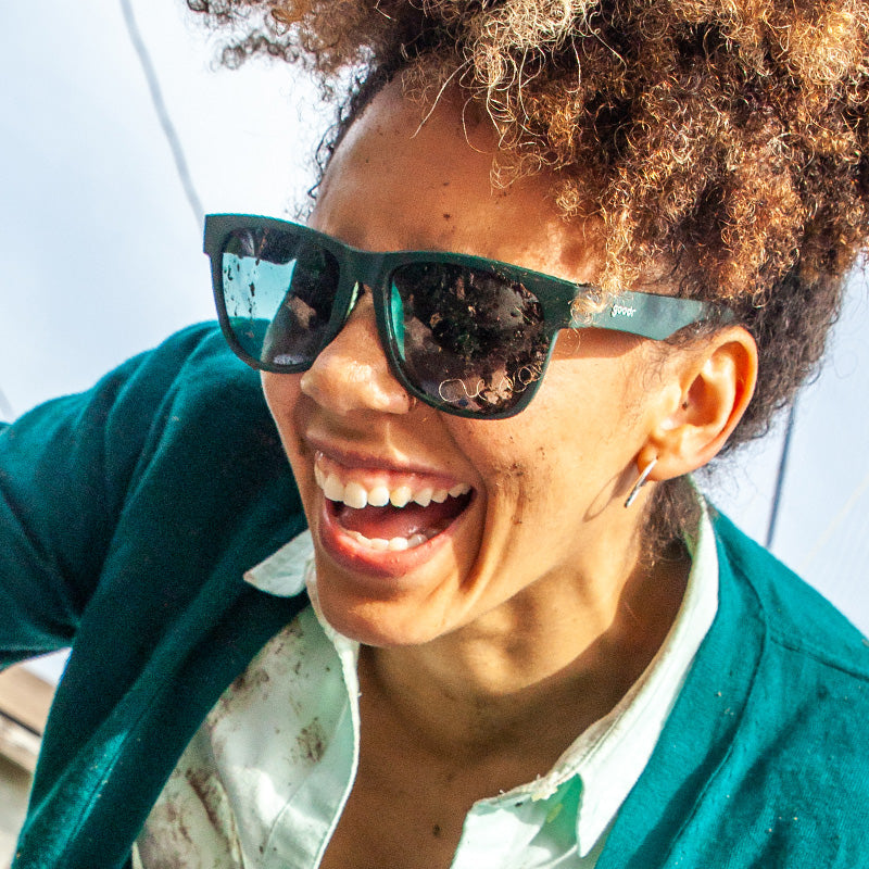 A woman shown smiling/laughing, wearing square sunglasses with teal green frames and a matching cardigan.